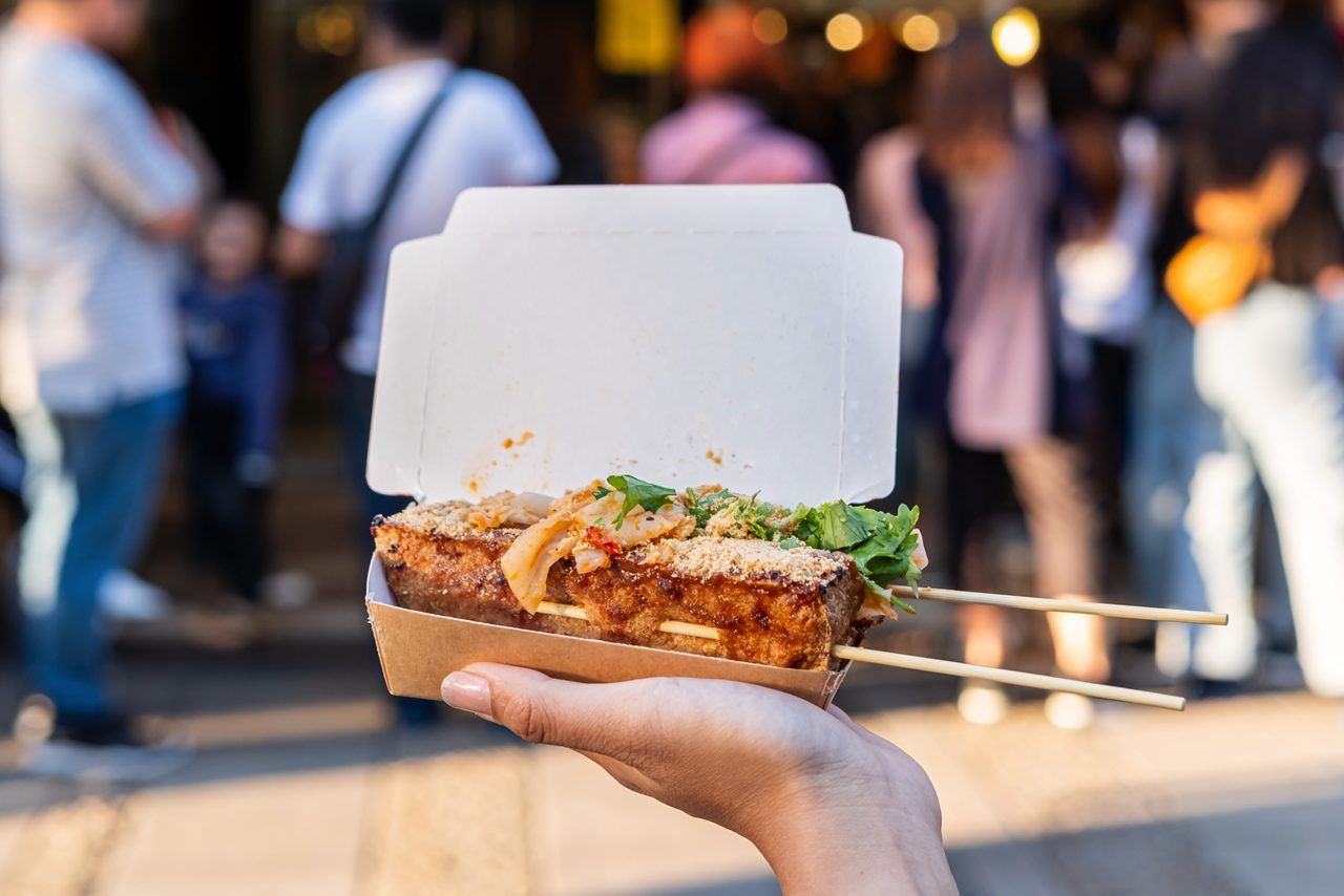 Close-up of grilled stinky tofu on skewers served in a paper tray, topped with kimchi and herbs, held by a hand in front of a bustling night market crowd in Taipei.