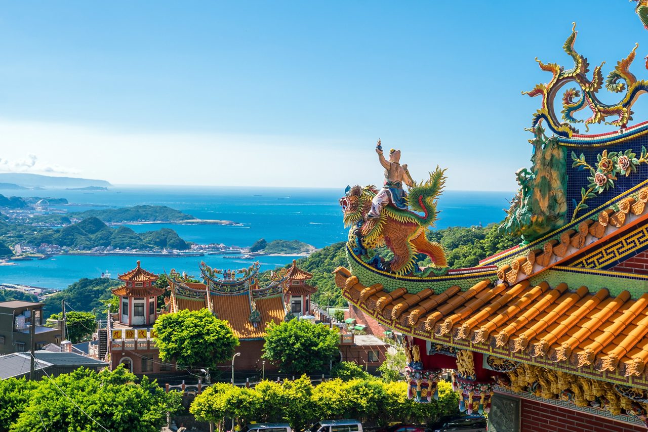 Colorful temple rooftop in Taipei, Taiwan adorned with intricate dragon and deity sculptures, overlooking a vibrant coastal landscape with deep blue ocean, green hills and scattered buildings below.