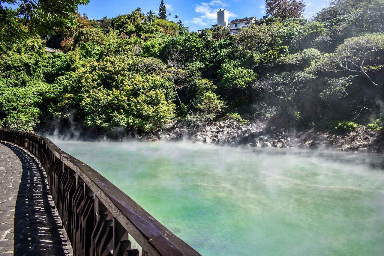 Steaming green hot spring pool surrounded by lush vegetation and rocks, with a shaded walkway along the water's edge in Yangmingshan National Park near Taipei, Taiwan.