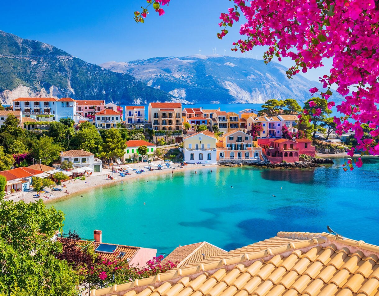 A colorful seaside village with pastel houses, pink bougainvillea and a sheltered turquoise bay surrounded by green hills.