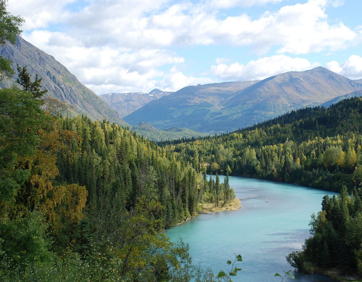 Aerial view of a turquoise river flowing through evergreen forest with snow-capped mountains in the background under partly cloudy skies