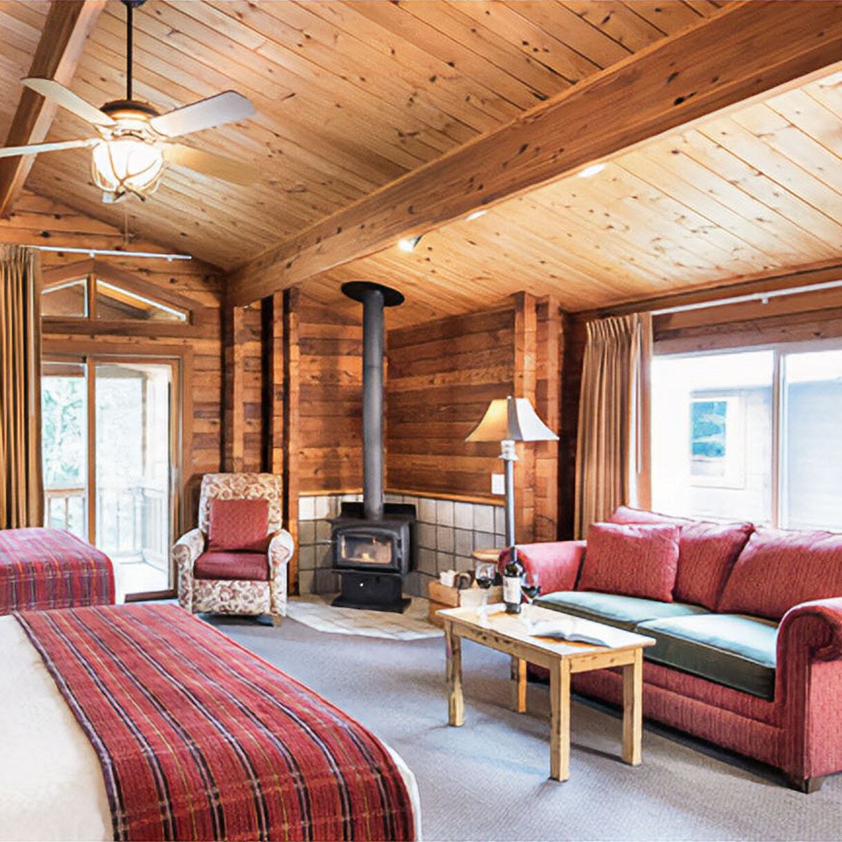 Rustic lodge room interior with wood-paneled walls, wood-burning stove, comfortable seating with coral-colored pillows, table lamp, and window overlooking water view