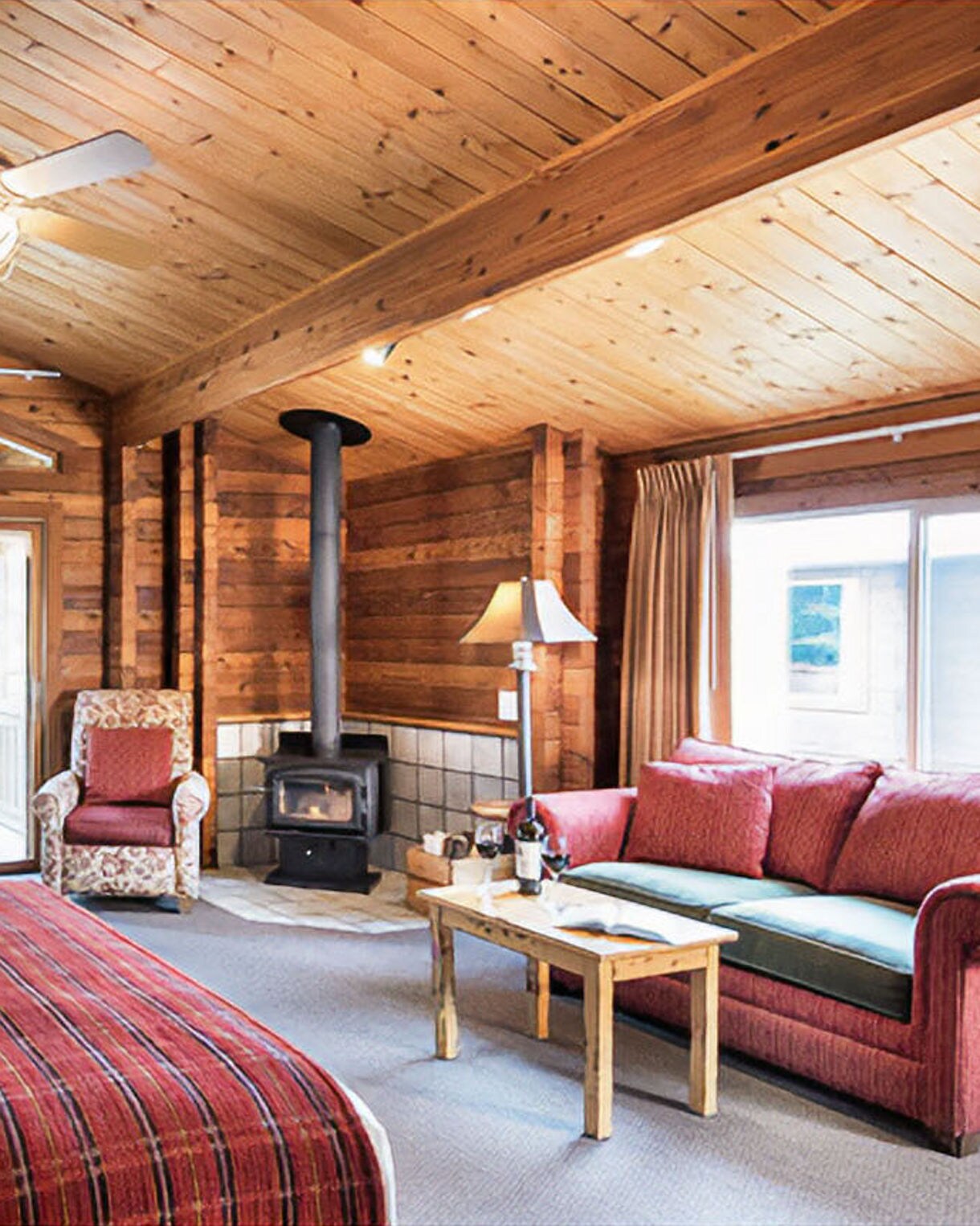 Rustic lodge room interior with wood-paneled walls, wood-burning stove, comfortable seating with coral-colored pillows, table lamp, and window overlooking water view