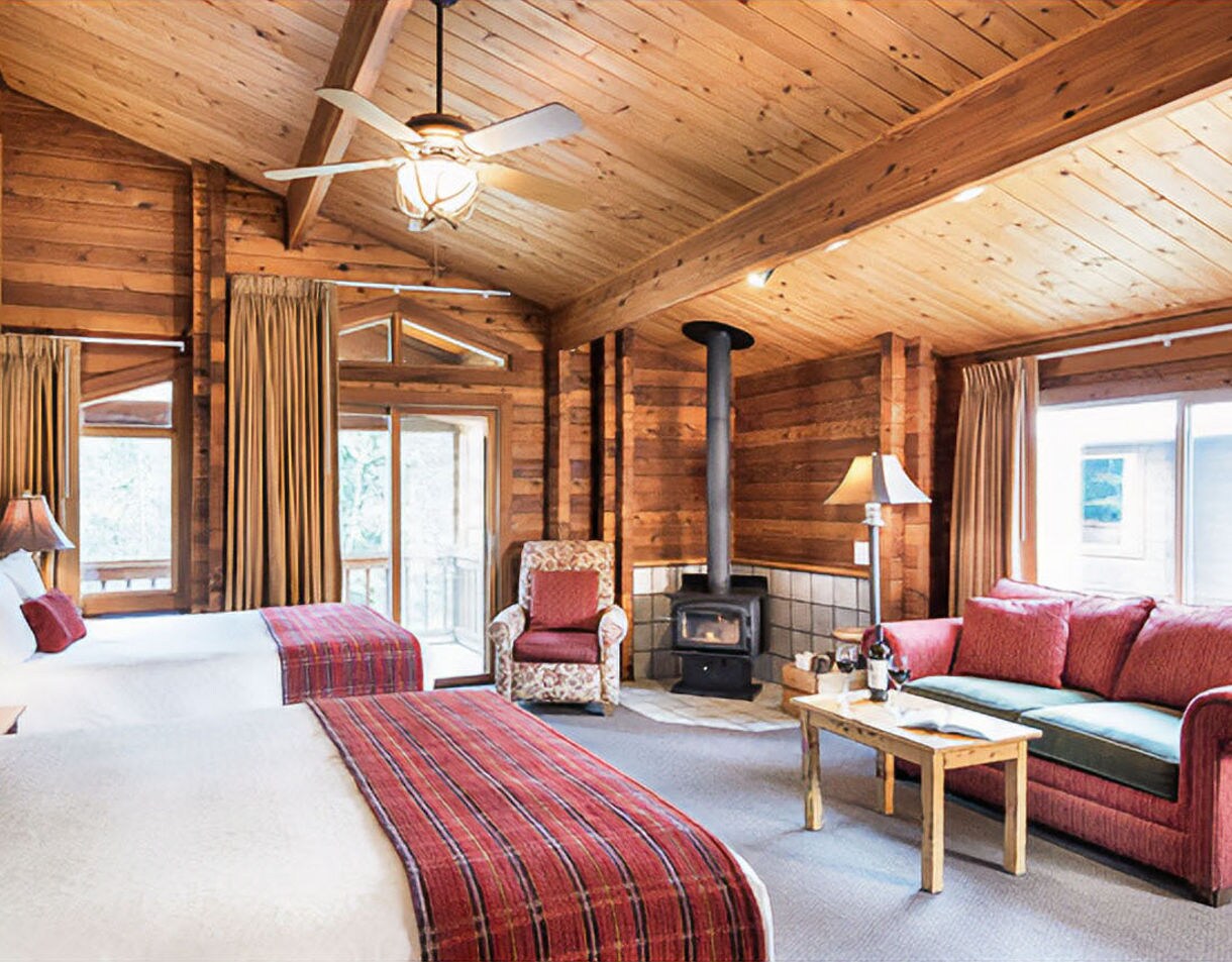 Rustic lodge room interior with wood-paneled walls, wood-burning stove, comfortable seating with coral-colored pillows, table lamp, and window overlooking water view