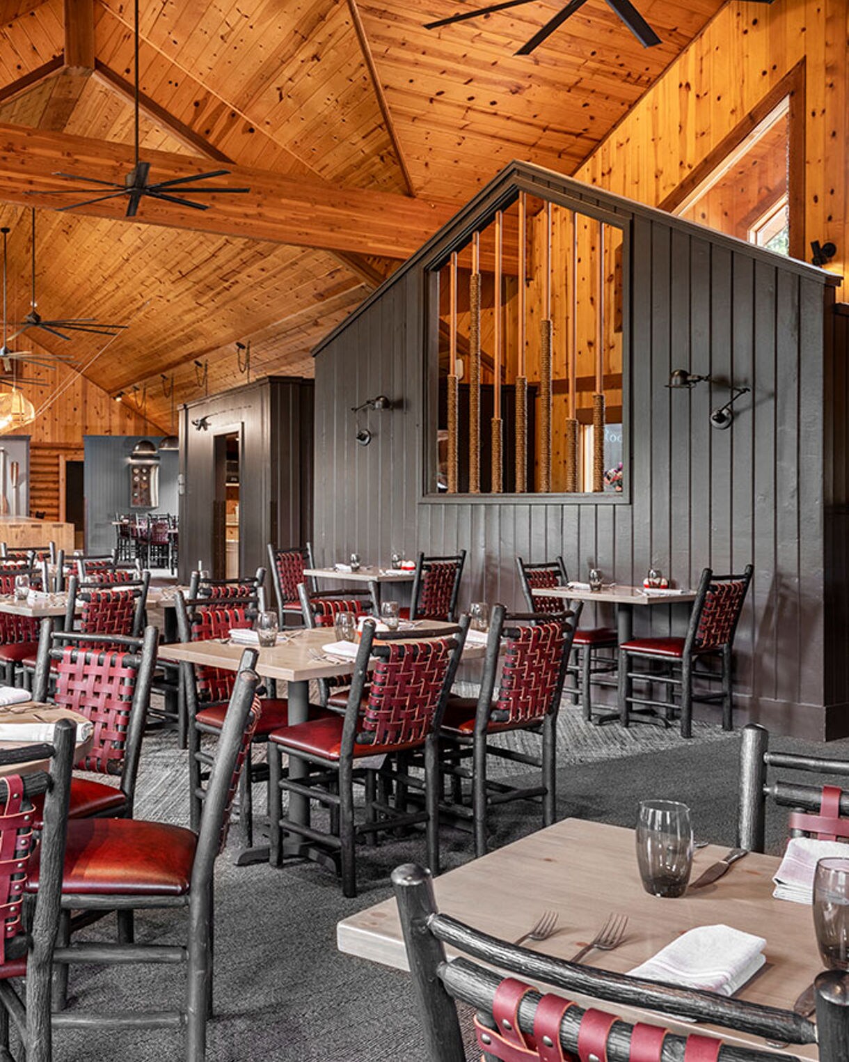 Spacious restaurant dining area features wooden walls, vaulted ceilings, and large windows letting in natural light. Tables are set with glassware, napkins, and utensils, while red woven chairs add a pop of color. The ambiance is warm and inviting, with rustic decor elements and a modern lodge style. No visible people or text are present in the image.
