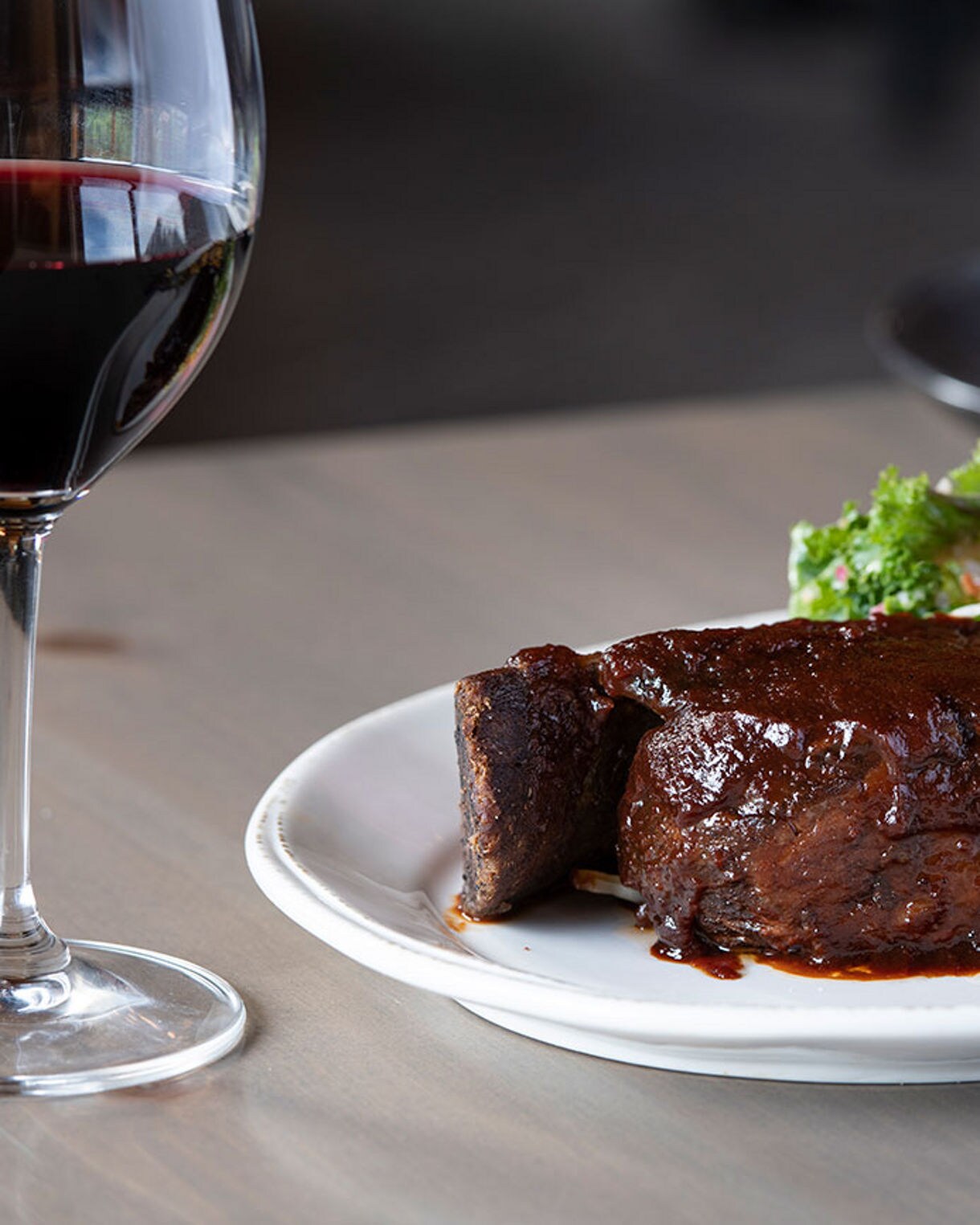 A succulent steak glazed with sauce is served alongside grilled vegetables and bread on a white plate. A glass of red wine sits nearby, enhancing the elegant dining atmosphere. The background features another dish, adding depth to the restaurant setting. The overall mood is refined and inviting, with a focus on rich, earthy tones.