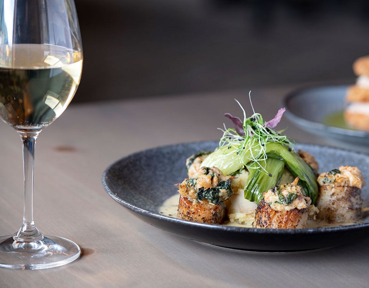 A plated gourmet seafood entrée is artfully presented with leafy greens and microgreens, accompanied by a glass of white wine. The setting is a modern restaurant table with soft, natural lighting. The background features another plated appetizer, adding to the upscale dining atmosphere. The overall mood is elegant and inviting, with a neutral color palette and refined presentation.