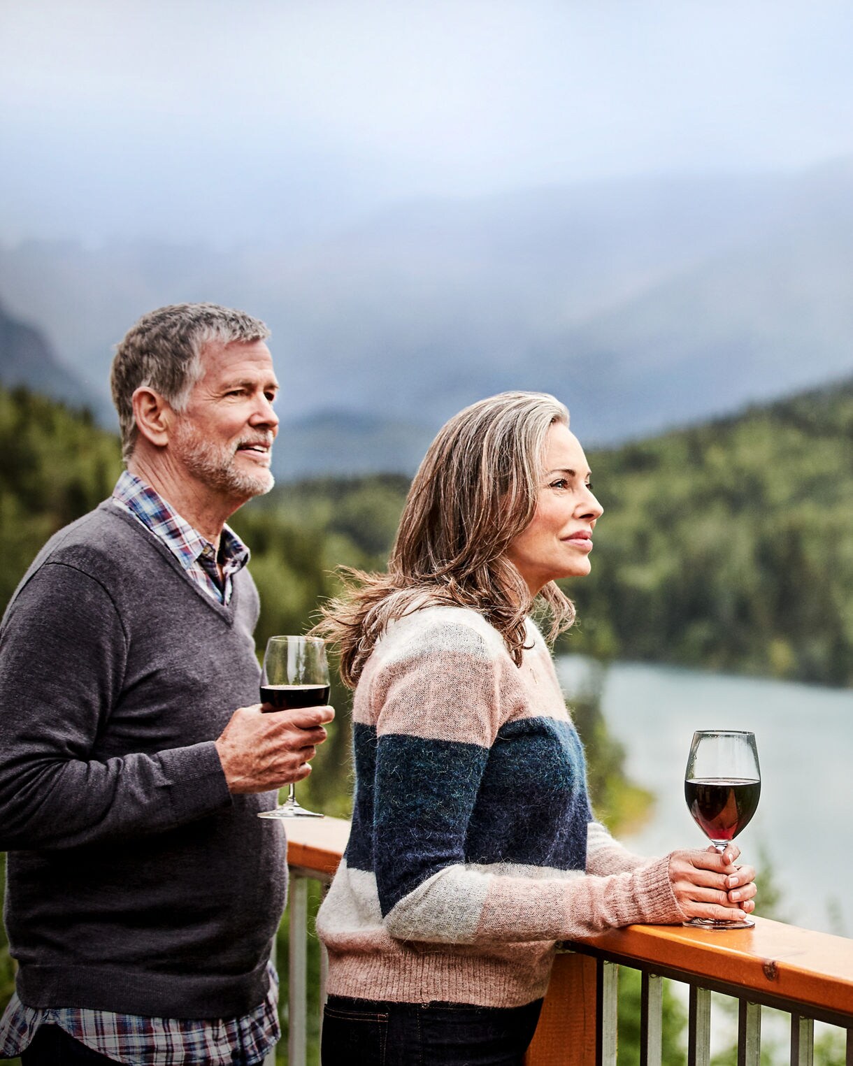 Couple standing on wooden deck holding wine glasses, overlooking a mountain lake surrounded by forest in misty conditions