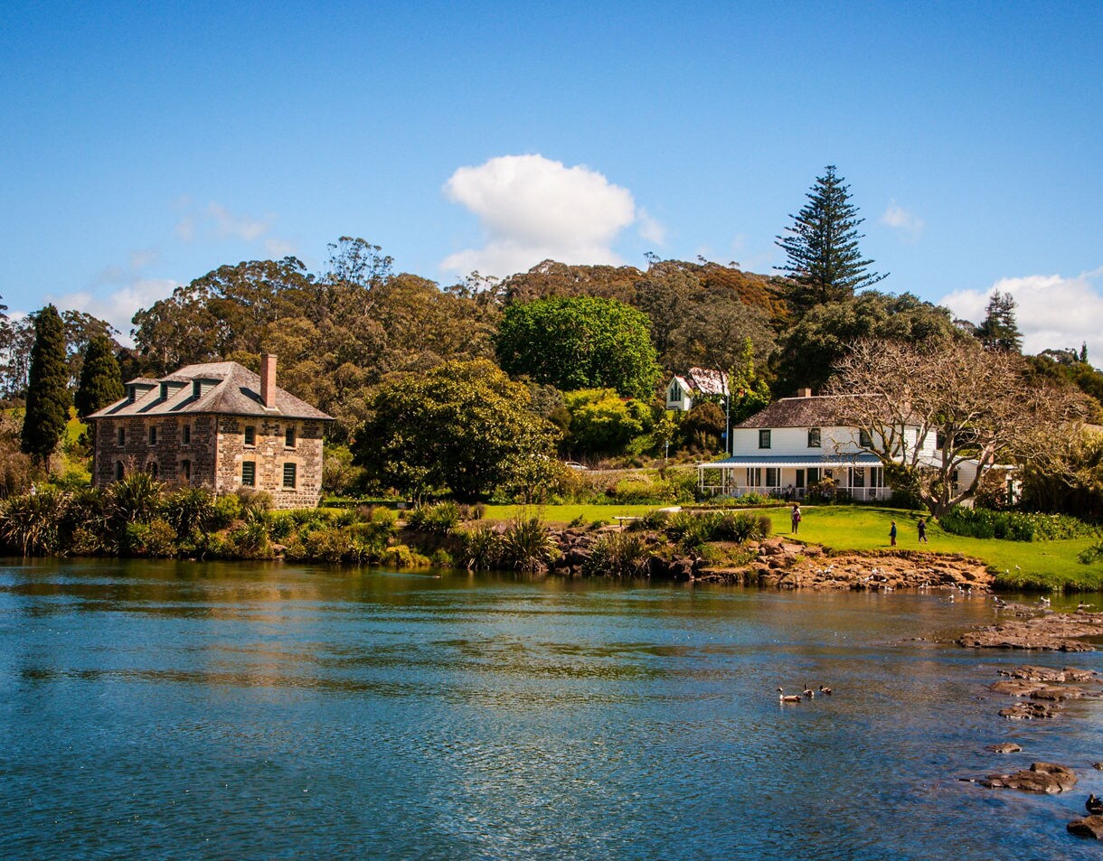 Historic stone store and colonial house beside a calm river in Kerikeri, New Zealand, surrounded by trees and bright green grass under a clear blue sky.