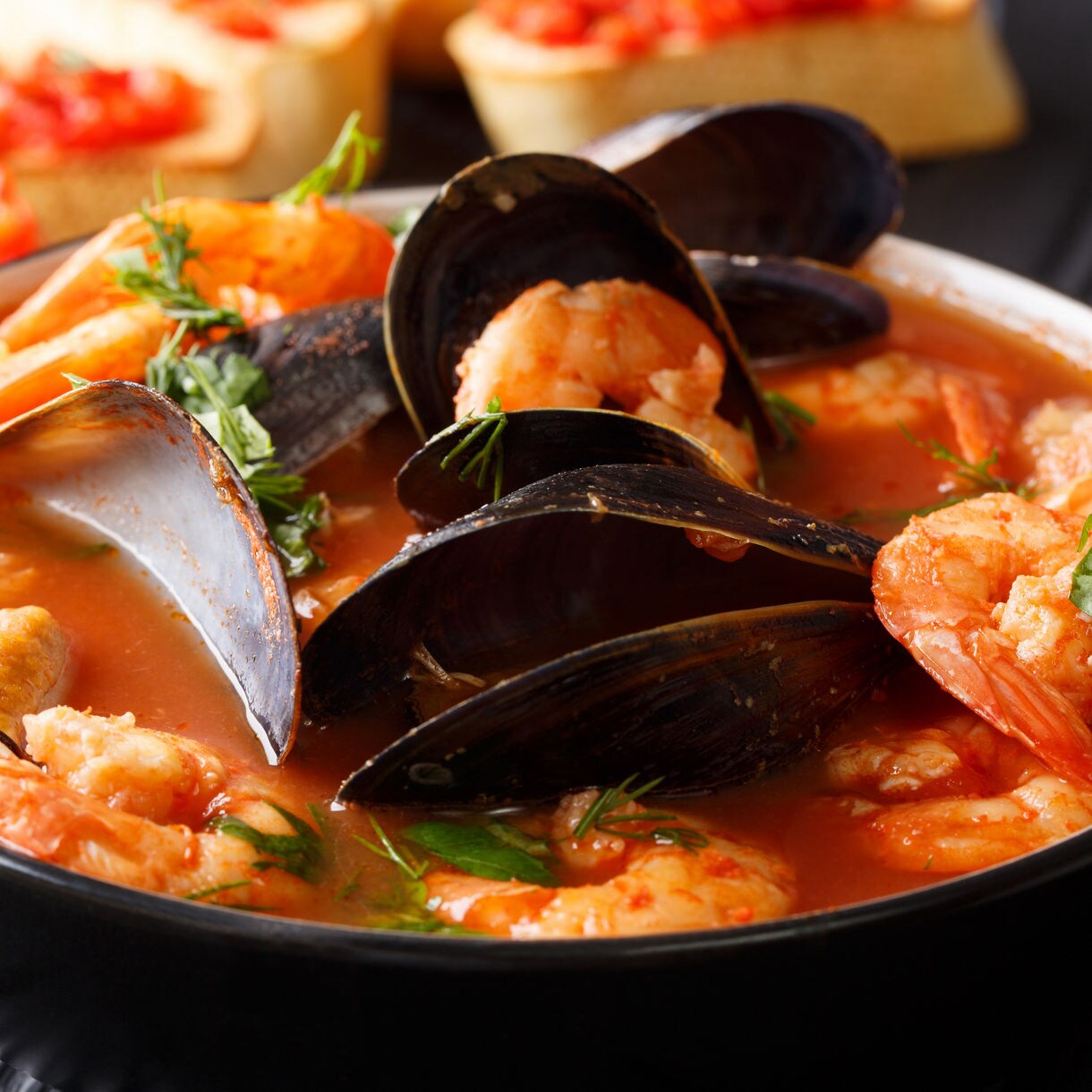A bowl of bouillabaisse featuring shrimp, mussels and salmon, garnished with fresh herbs, alongside toasted bread with a tomato topping.