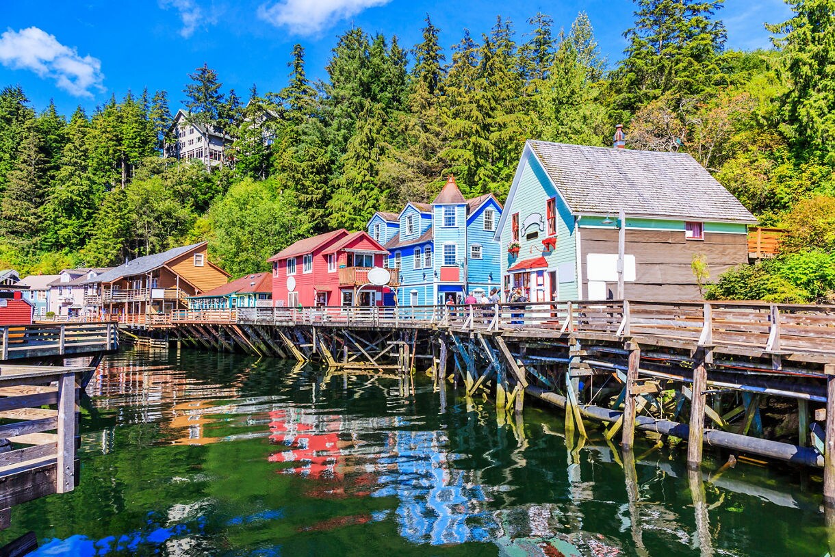 A vibrant scene of Creek Street in Ketchikan, Alaska, featuring colorful historic buildings on wooden stilts above reflective water, framed by dense green trees.