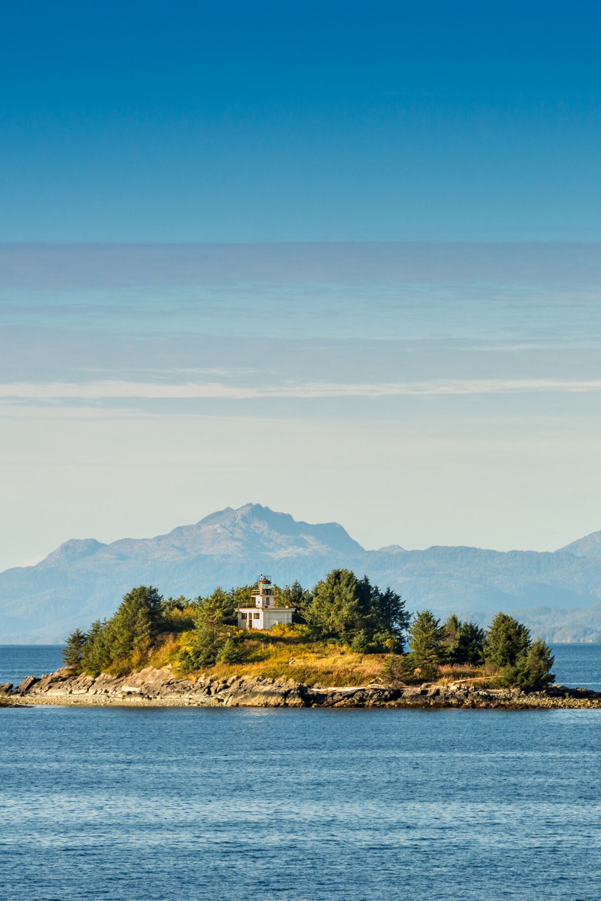 A small lighthouse perched on an island, surrounded by trees and calm water, with distant mountain ranges under a clear blue sky.