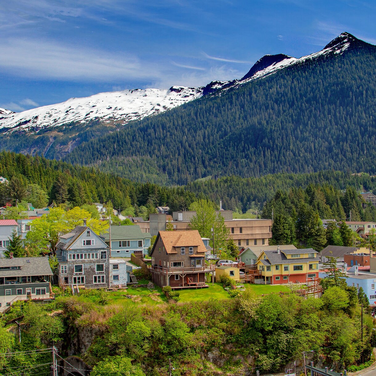 A scenic view of Ketchikan with colorful houses in the foreground and snow-capped mountains rising in the background, framed by lush green forests.