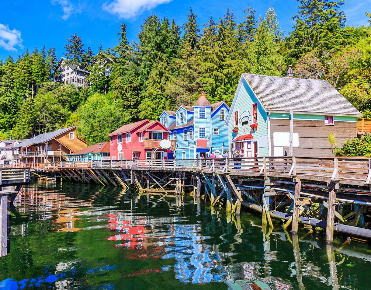 Brightly colored buildings line the wooden dock in Ketchikan, reflecting in the calm waters under a blue sky, with lush greenery in the background.