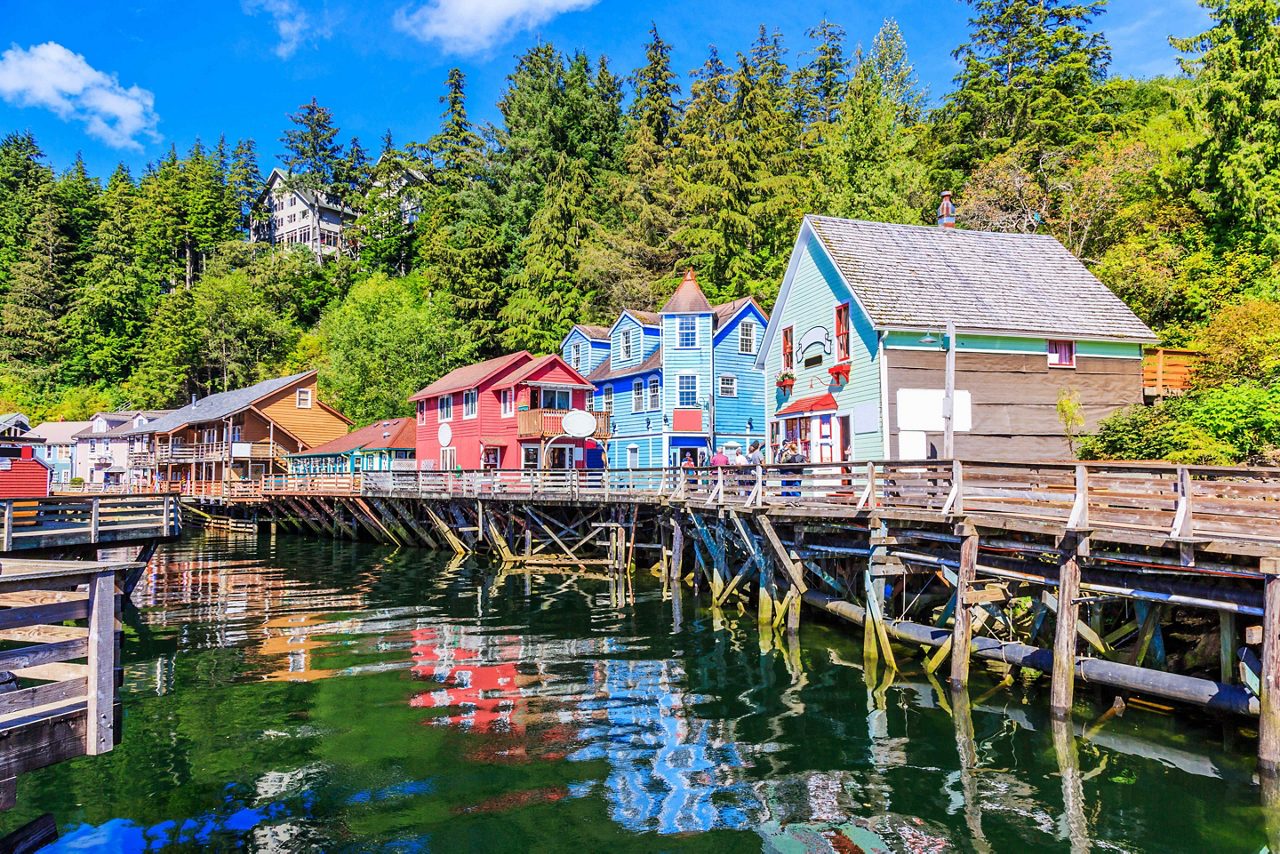 Brightly colored buildings line the wooden dock in Ketchikan, reflecting in the calm waters under a blue sky, with lush greenery in the background.