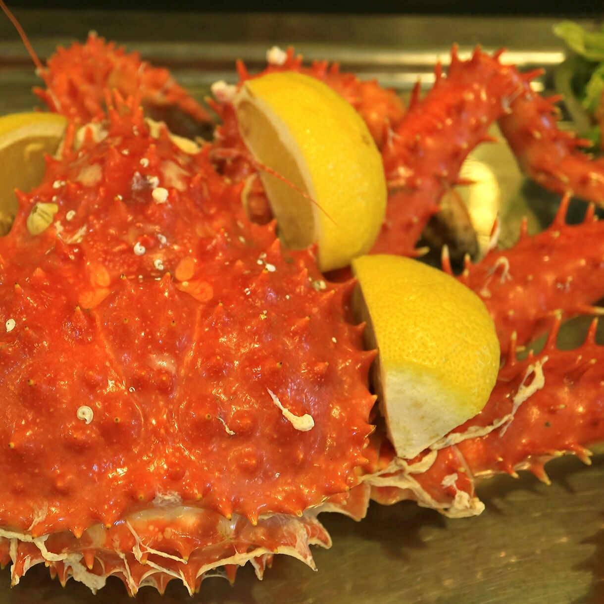 Close-up of a cooked red king crab topped with lemon wedges, displayed on a metal serving tray beside a portion of yellow rice.
