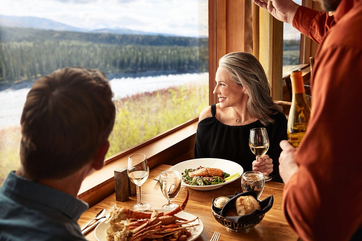 Three adults are seated at a wooden table by a large window overlooking a river and forested hills. The table is set with plates of seafood, salad, bread, and glasses of white wine, creating a relaxed and elegant dining atmosphere. Warm natural light fills the space, highlighting the cozy and inviting mood. The setting suggests a casual yet refined meal with a beautiful outdoor view.