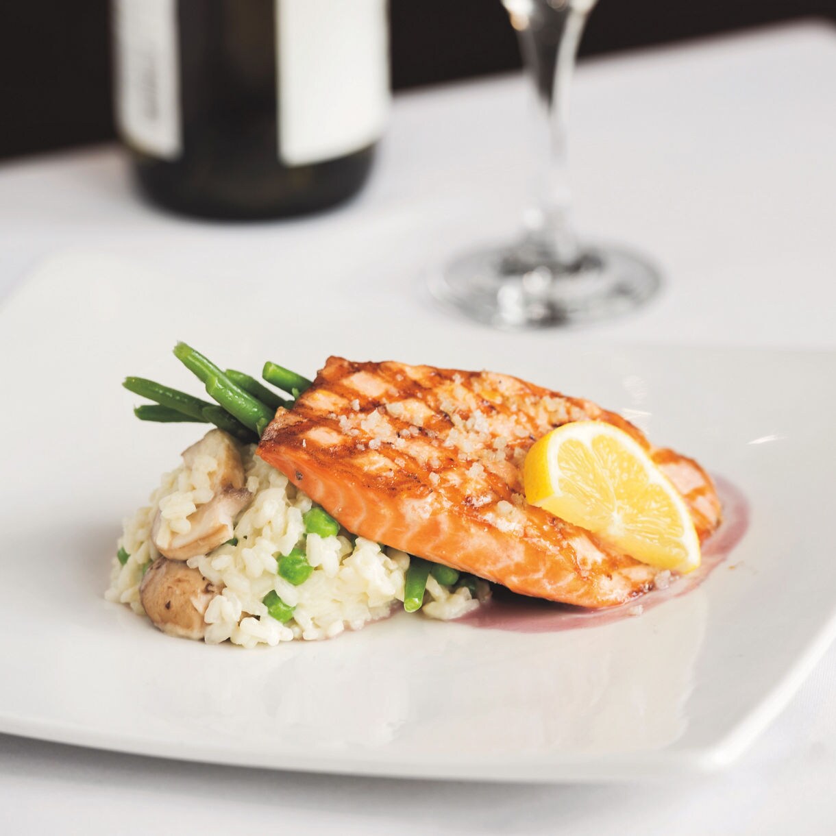 A grilled salmon fillet is elegantly plated atop a serving of creamy risotto with green beans and mushrooms. A fresh lemon wedge garnishes the fish, adding a pop of color and freshness. The dish is presented on a modern white plate, with a blurred wine bottle and glass in the background, suggesting a fine dining setting. The overall mood is sophisticated and appetizing, with a neutral and warm palette.