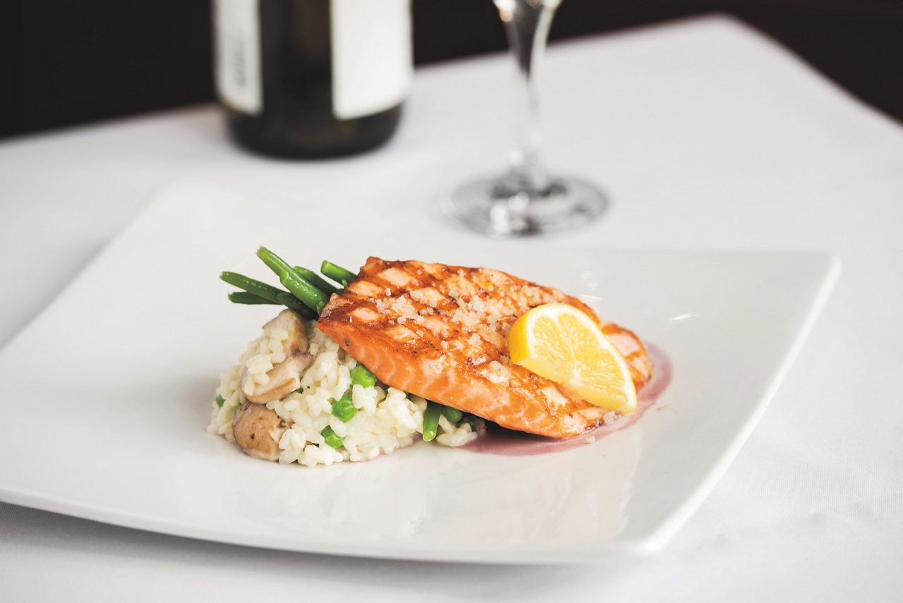 A grilled salmon fillet is elegantly plated atop a serving of creamy risotto with green beans and mushrooms. A fresh lemon wedge garnishes the fish, adding a pop of color and freshness. The dish is presented on a modern white plate, with a blurred wine bottle and glass in the background, suggesting a fine dining setting. The overall mood is sophisticated and appetizing, with a neutral and warm palette.