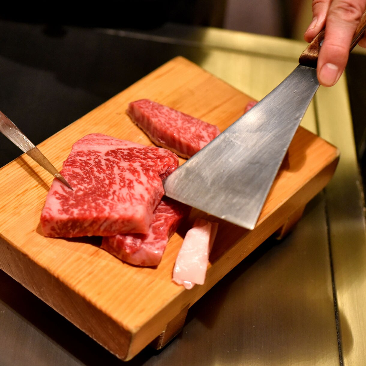 Close-up of raw Kobe beef being sliced on a wooden board with metal utensils at a teppanyaki grill.