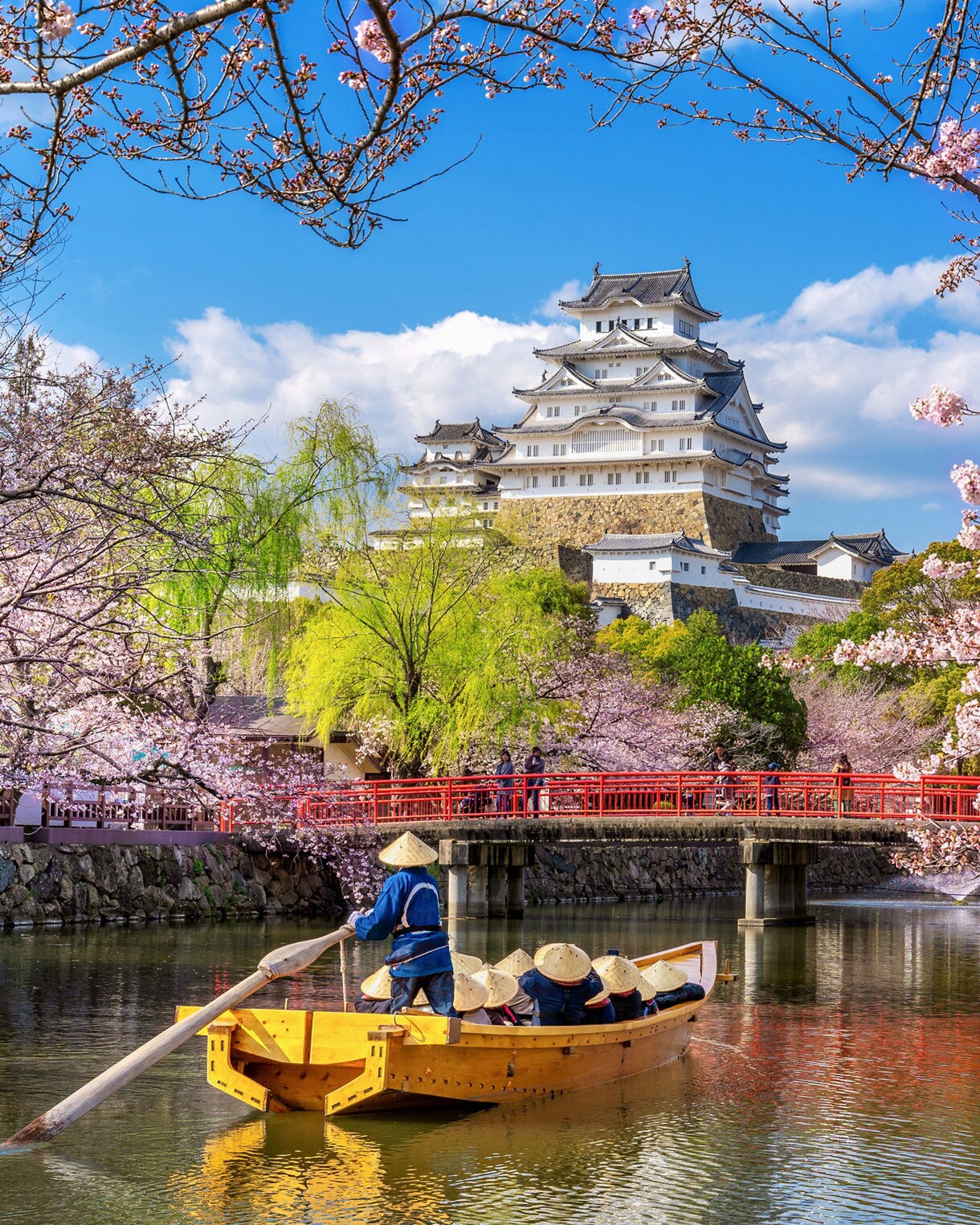 raditional wooden boat carrying passengers along a stone-walled waterway under an archway of pink cherry blossoms, with a multi-tiered white Japanese castle and red bridge visible in the background.