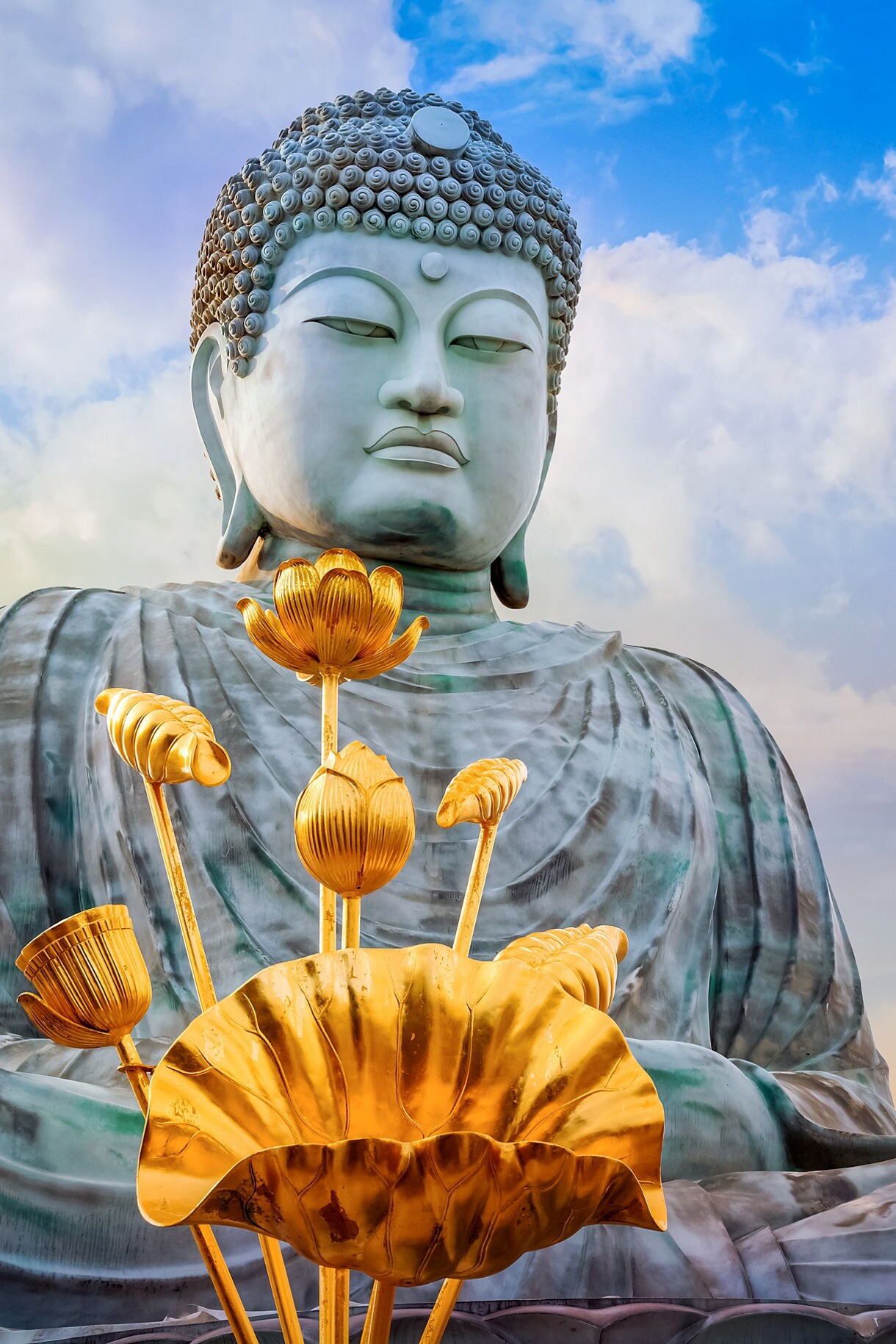Close-up of the Hyogo Daibutsu Buddha statue in Kobe, Japan, with golden lotus flowers in front and a vibrant sunset sky in the background.