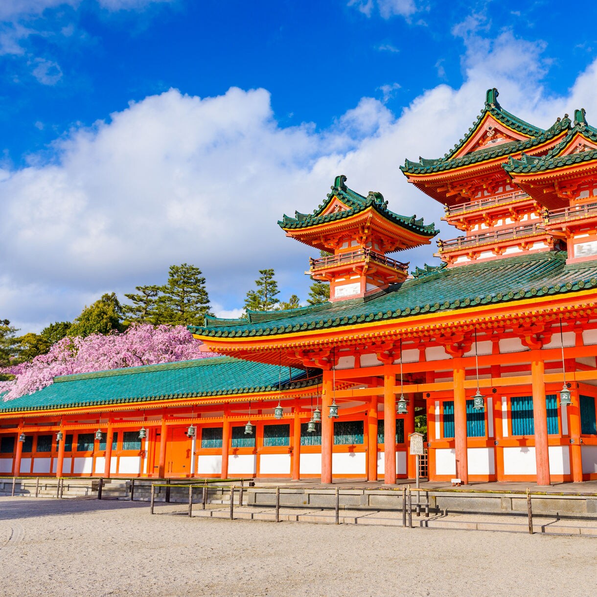 Bright red Heian Shrine in Kyoto, Japan with green-tiled roofs, cherry blossoms and blue sky in the background.