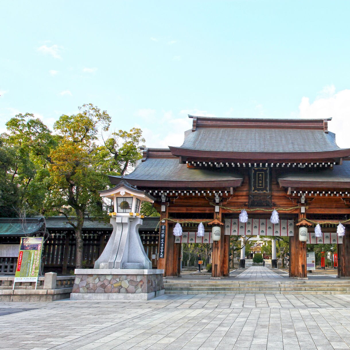 Traditional wooden torii gate of Minatogawa Shrine in Kobe, Japan, flanked by stone lanterns and trees.