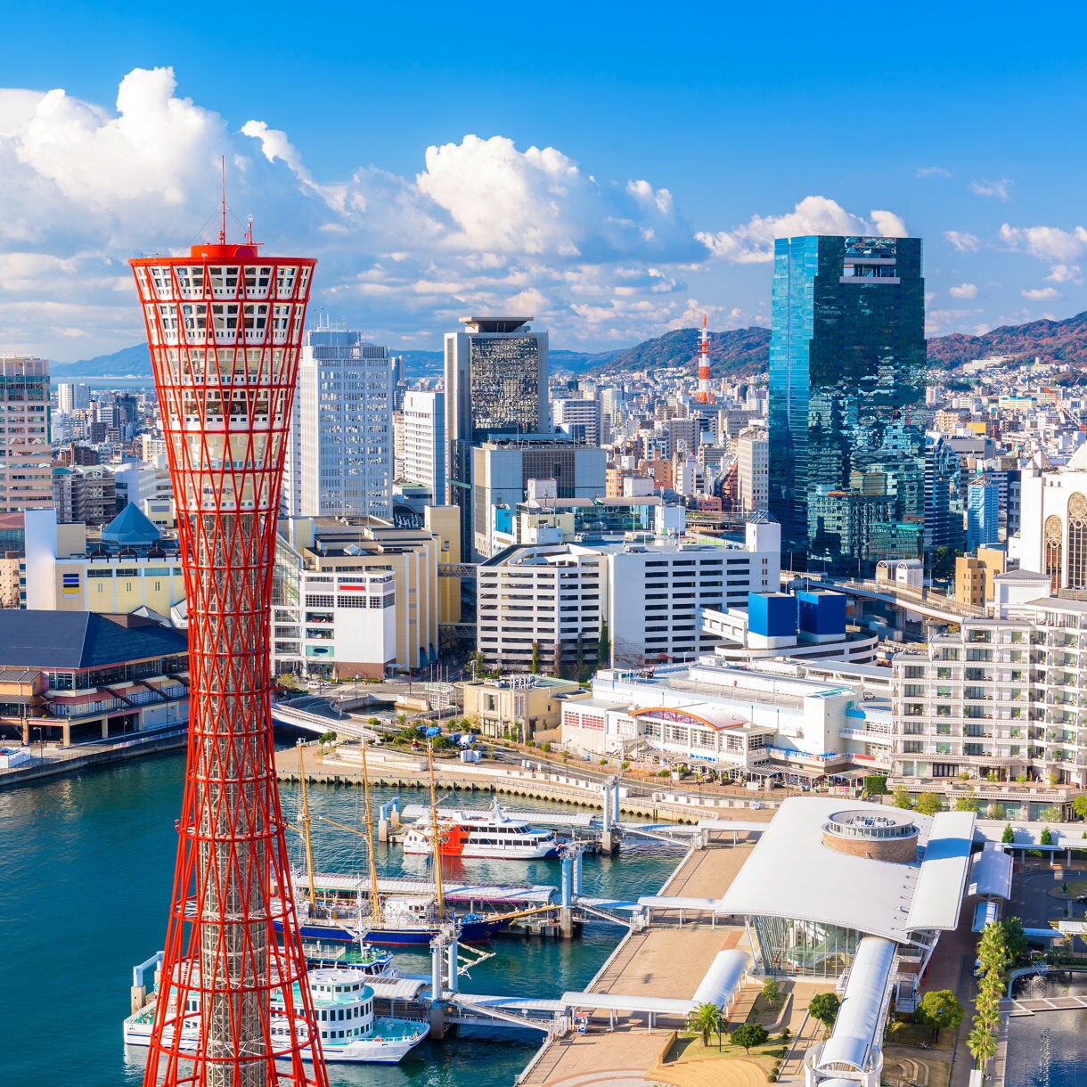 Kobe Port skyline featuring the red Kobe Port Tower, waterfront buildings, docked boats and surrounding cityscape under a bright blue sky.