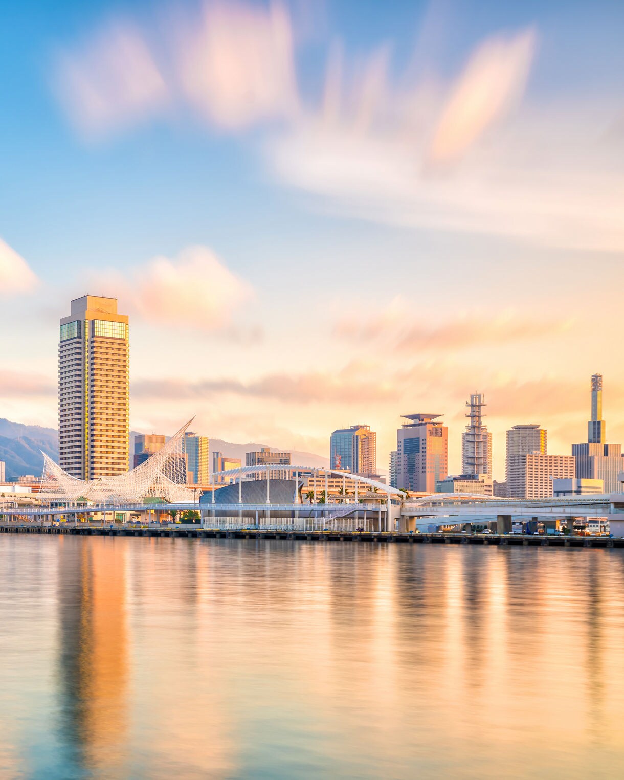 View of Kobe’s waterfront skyline at sunset with the red Port Tower, modern buildings and mountains in the background.