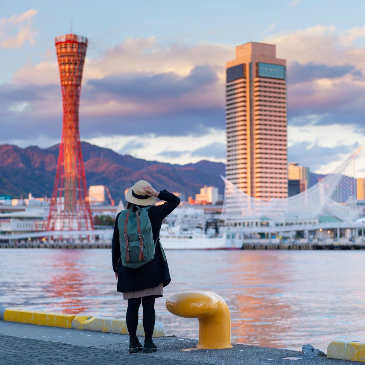A person wearing a backpack and hat stands near the waterfront in Kobe, Japan, gazing at the city skyline featuring the red Kobe Port Tower, contemporary buildings, and distant mountains under a colorful sky