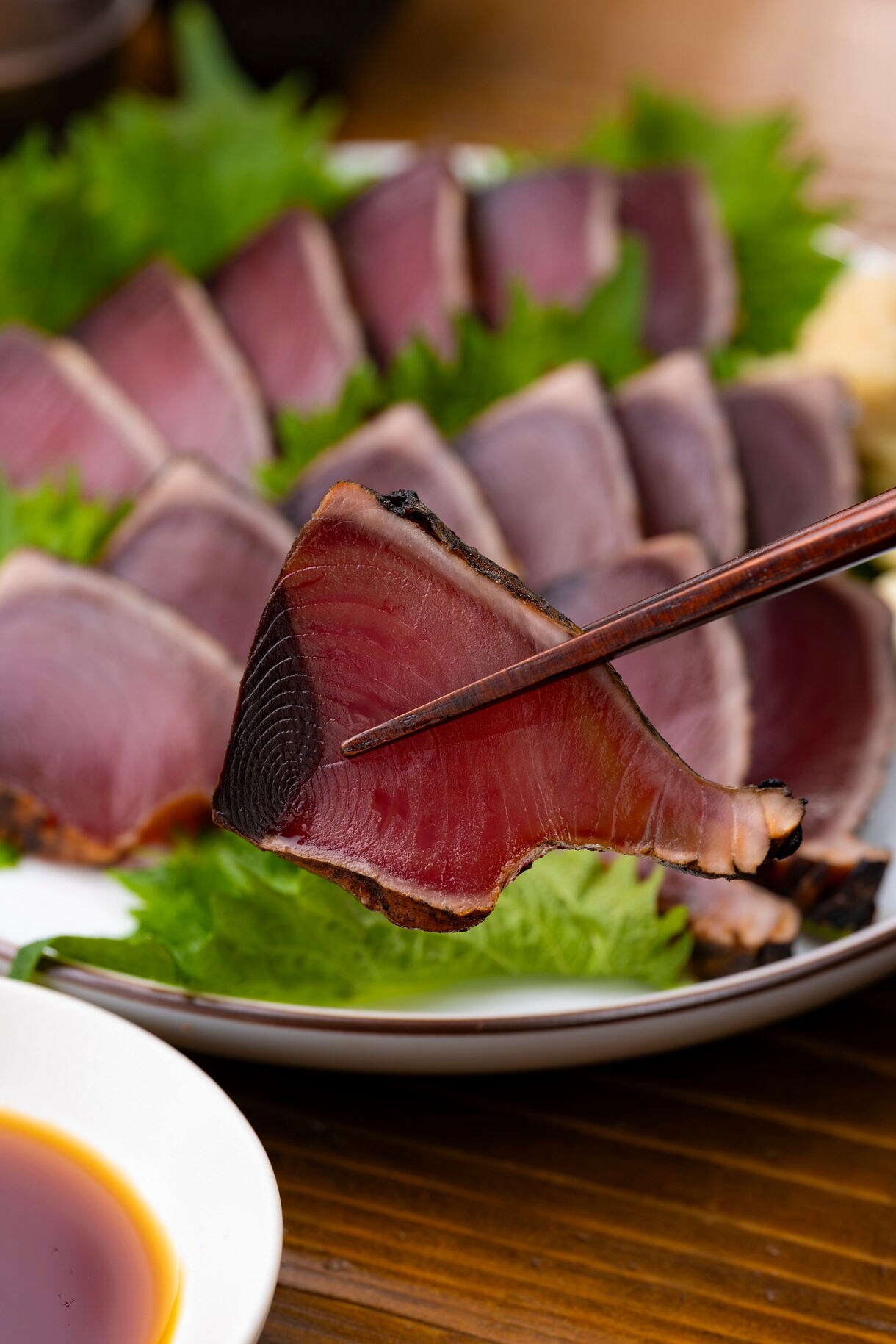 Close-up of a slice of seared bonito held with chopsticks above a plate of sliced fish and green garnish.