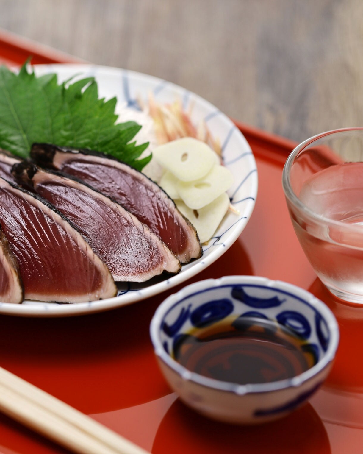 Plate of seared bonito slices with lime, garlic and garnish, served on a red tray with dipping sauce and sake.