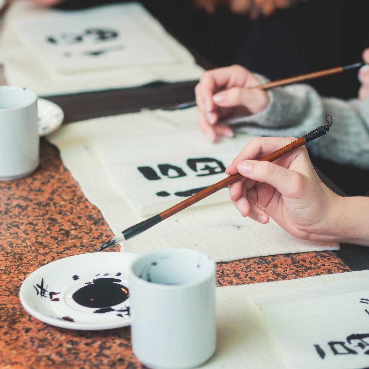 Hands holding calligraphy brushes writing Japanese characters on white paper with ink dishes and cups on the table.