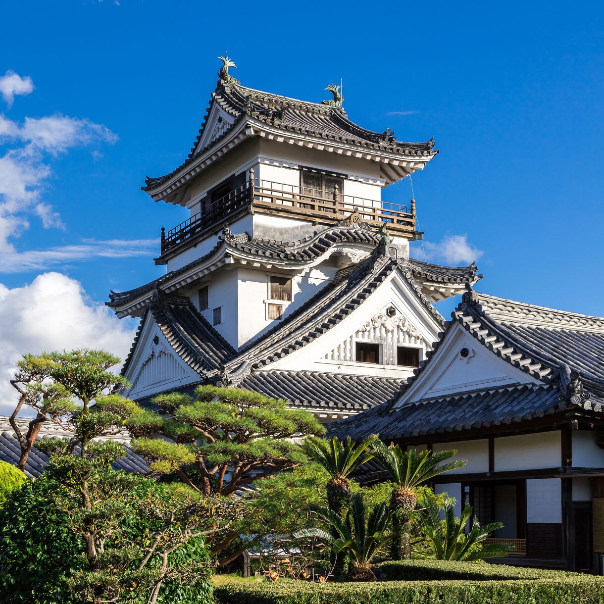 Traditional Japanese castle with curved tile roofs and white walls, framed by manicured trees and blue sky.