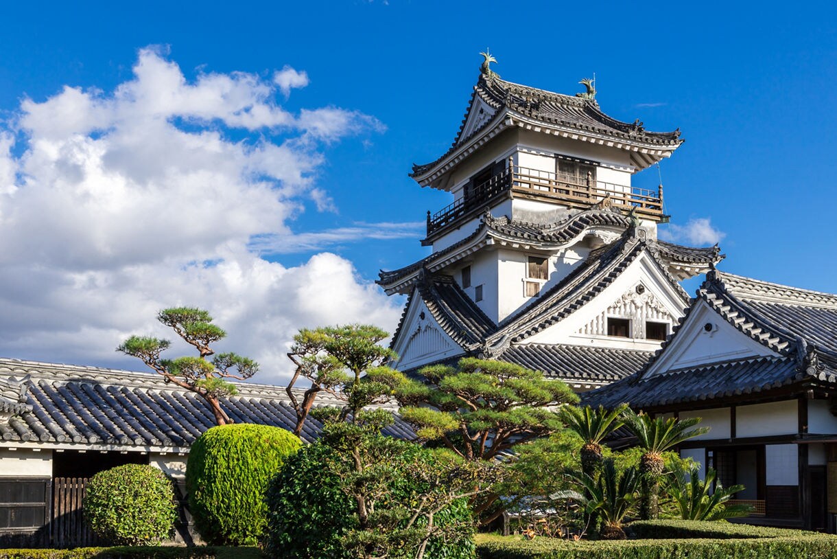 Traditional Japanese castle with curved tile roofs and white walls, framed by manicured trees and blue sky.