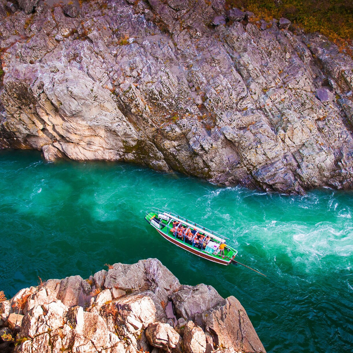 Boat with tourists gliding through the green waters of Oboke Gorge, surrounded by steep rocky cliffs.