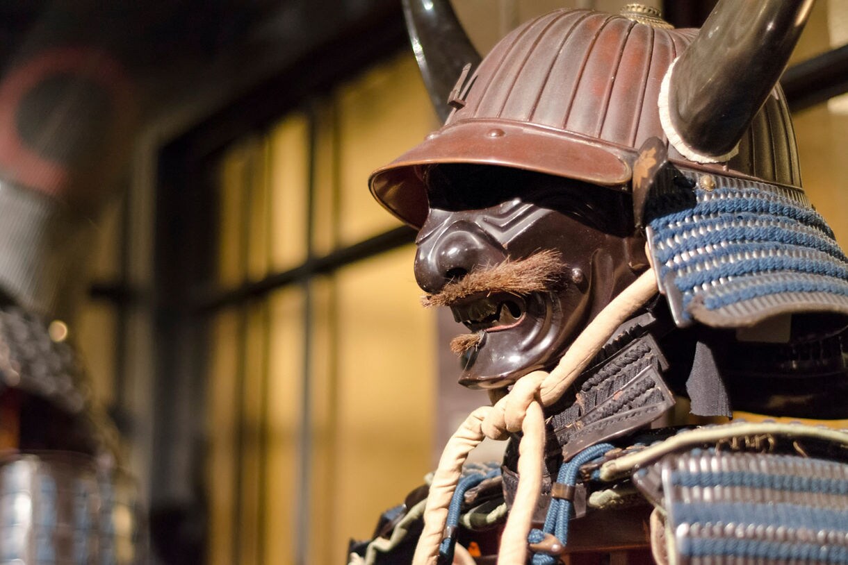 Close-up of traditional Japanese samurai armor featuring a horned helmet, mustached mask and shoulder guards.