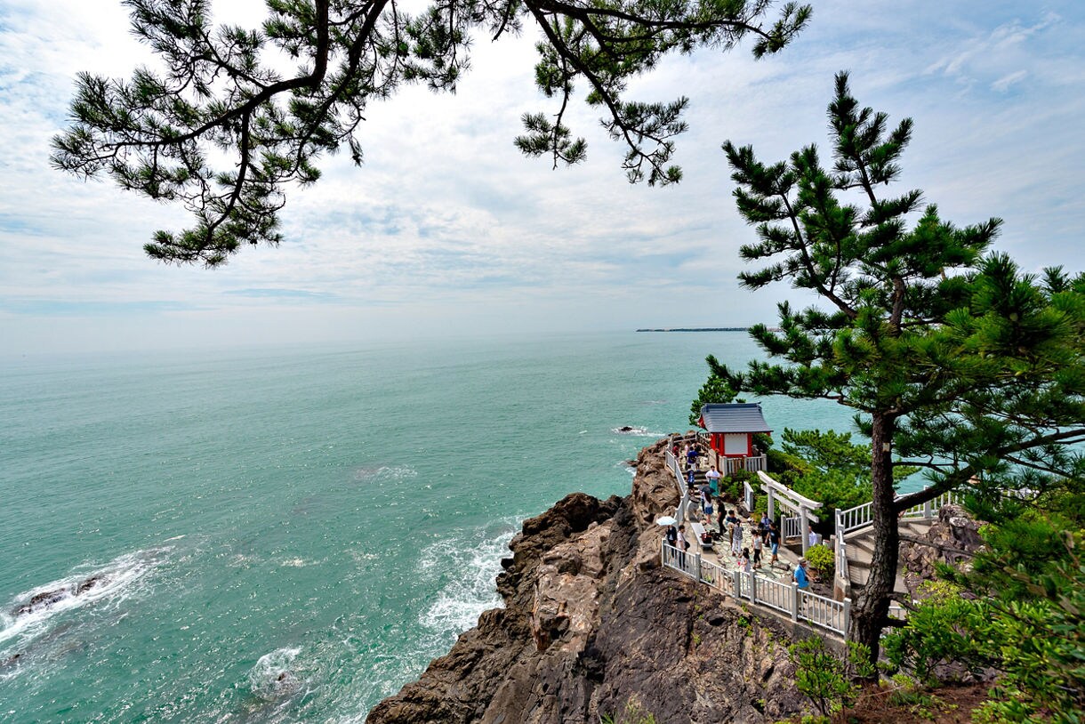 Tourists walking along a seaside cliff path leading to a small red and white shrine, with pine trees and ocean views.