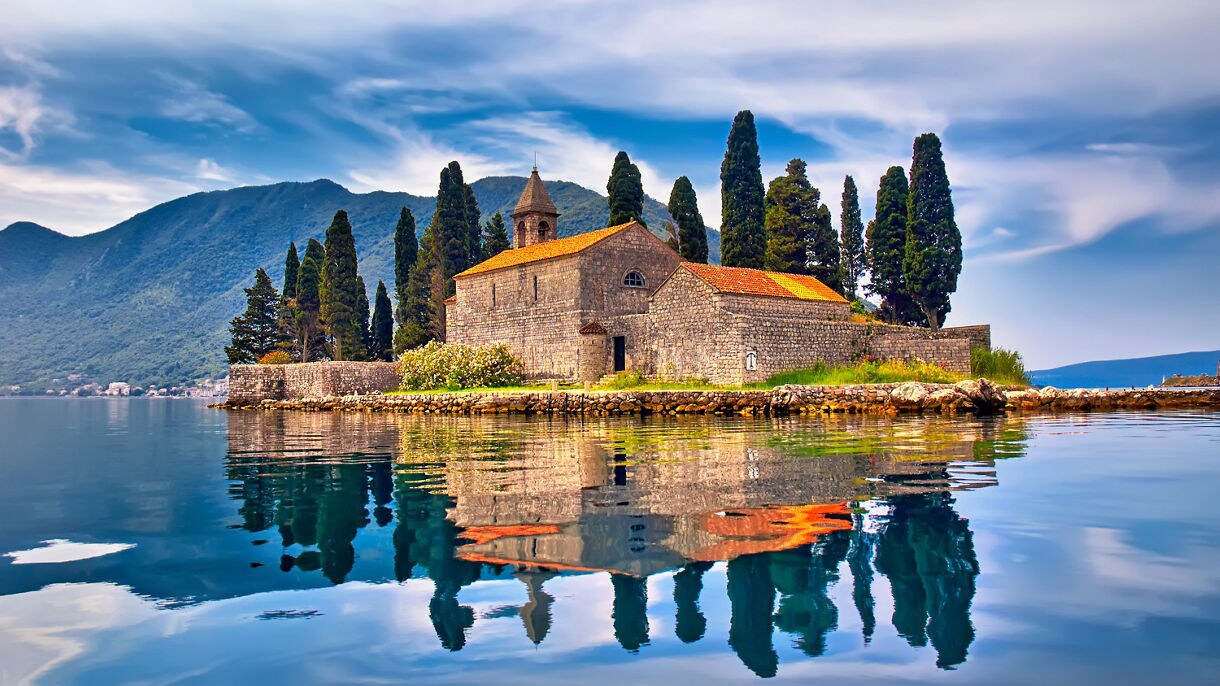 St. George Island in Montenegro, featuring an old stone church surrounded by cypress trees, reflected in the still waters with mountains in the background and a cloudy sky overhead.