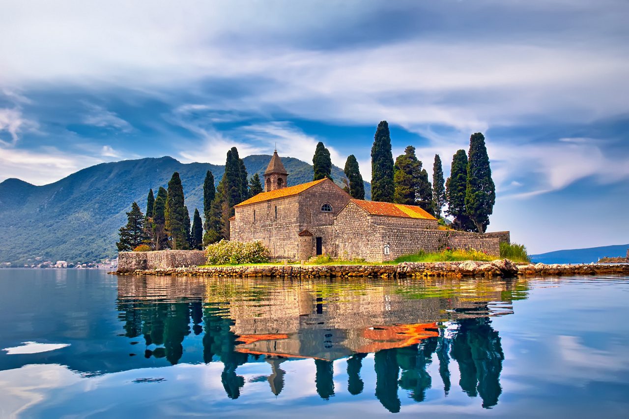 St. George Island in Montenegro, featuring an old stone church surrounded by cypress trees, reflected in the still waters with mountains in the background and a cloudy sky overhead.