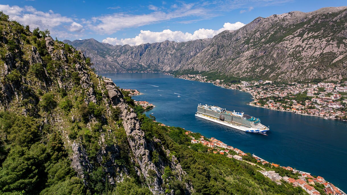 A cruise ship gliding through a narrow bay in Kotor, Montenegro, surrounded by steep green mountains, red‑roofed coastal towns, and bright blue water under a clear sky.