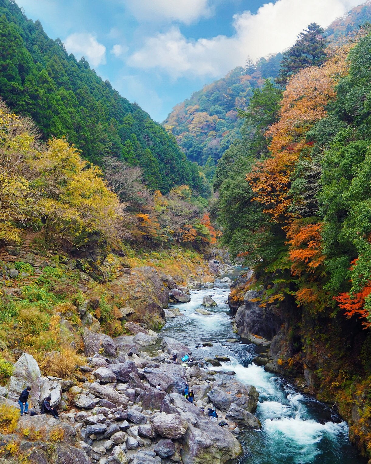 A winding river cuts through steep, rocky cliffs covered in autumn foliage and evergreen trees, with people exploring the boulders along Dorokyo Gorge.
