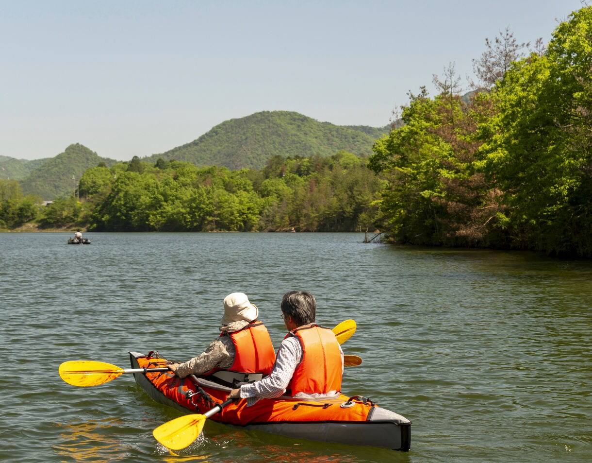 Two people wearing orange life vests paddle a red kayak through a calm forested lake surrounded by green hills under a clear sky.