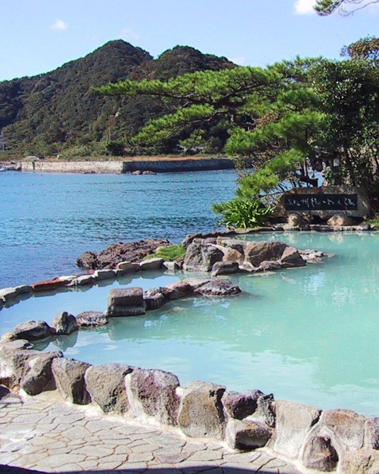Rock-lined outdoor hot spring pool with milky blue water sits beside a calm bay, surrounded by greenery and forested mountains in Nachikatsuura, Japan.