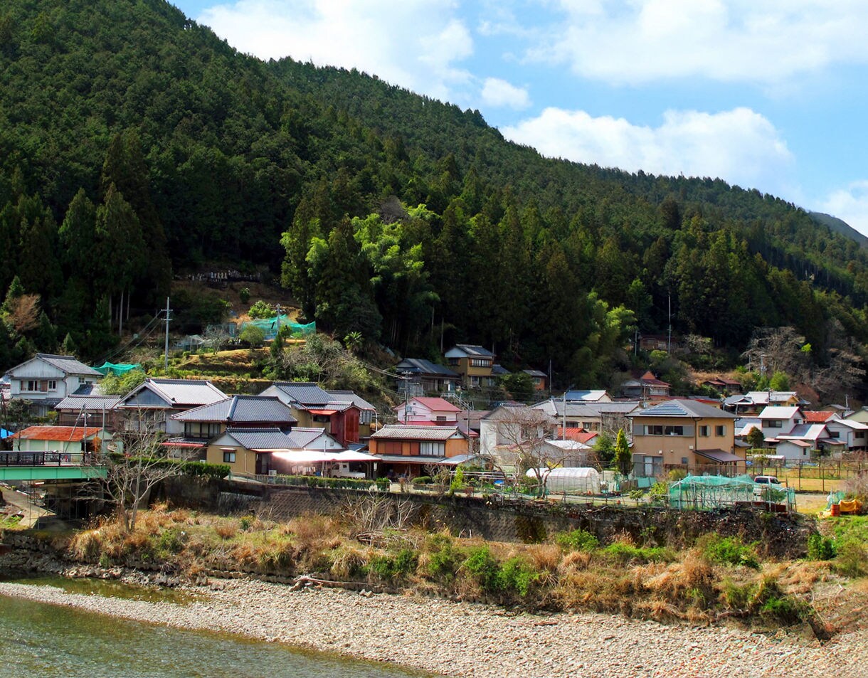 Traditional Japanese houses with tiled roofs sit at the base of a green forested mountain along a rocky riverbank in a rural Kumano town.