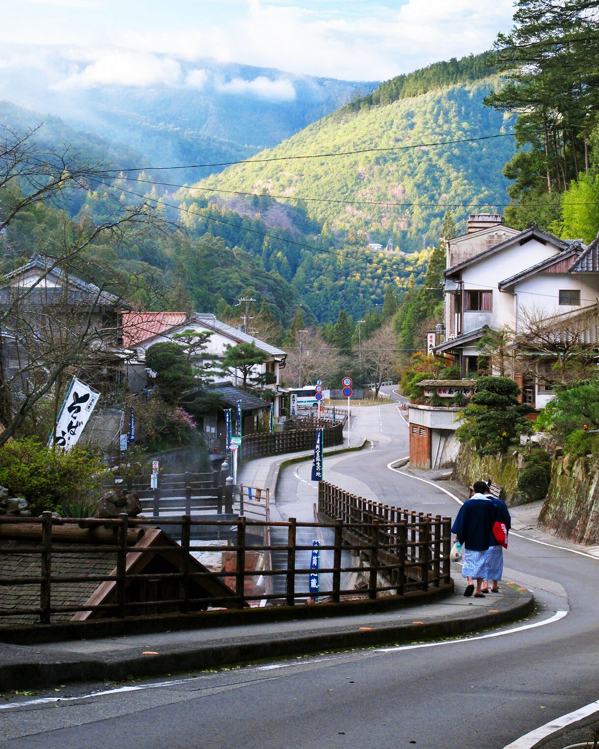 A traditional Japanese village with wooden houses and red tile roofs situated in a green mountain valley, with a person in traditional dress walking along a road beside a wooden railing.
