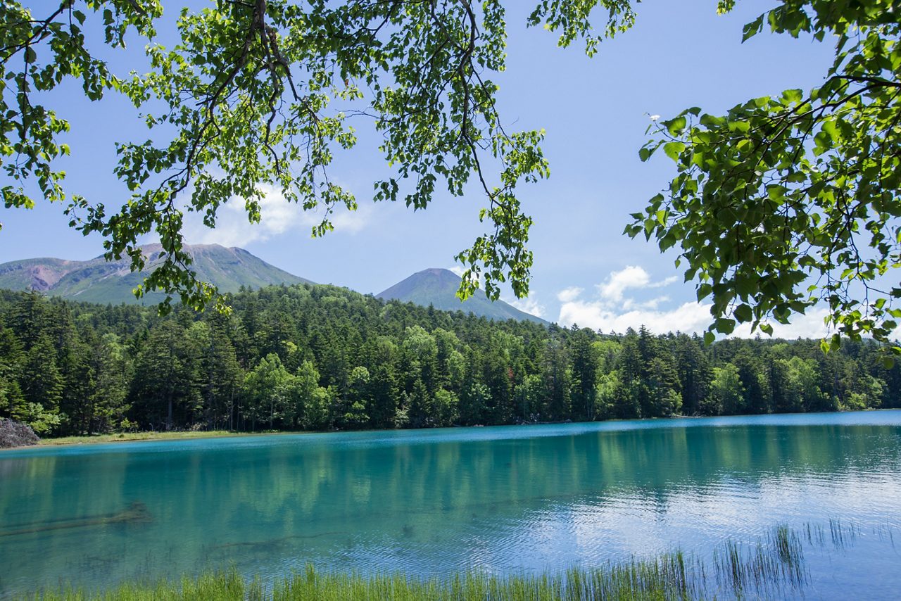 Clear blue Lake Akan bordered by lush green forest, with mountains in the background and tree branches framing the top.