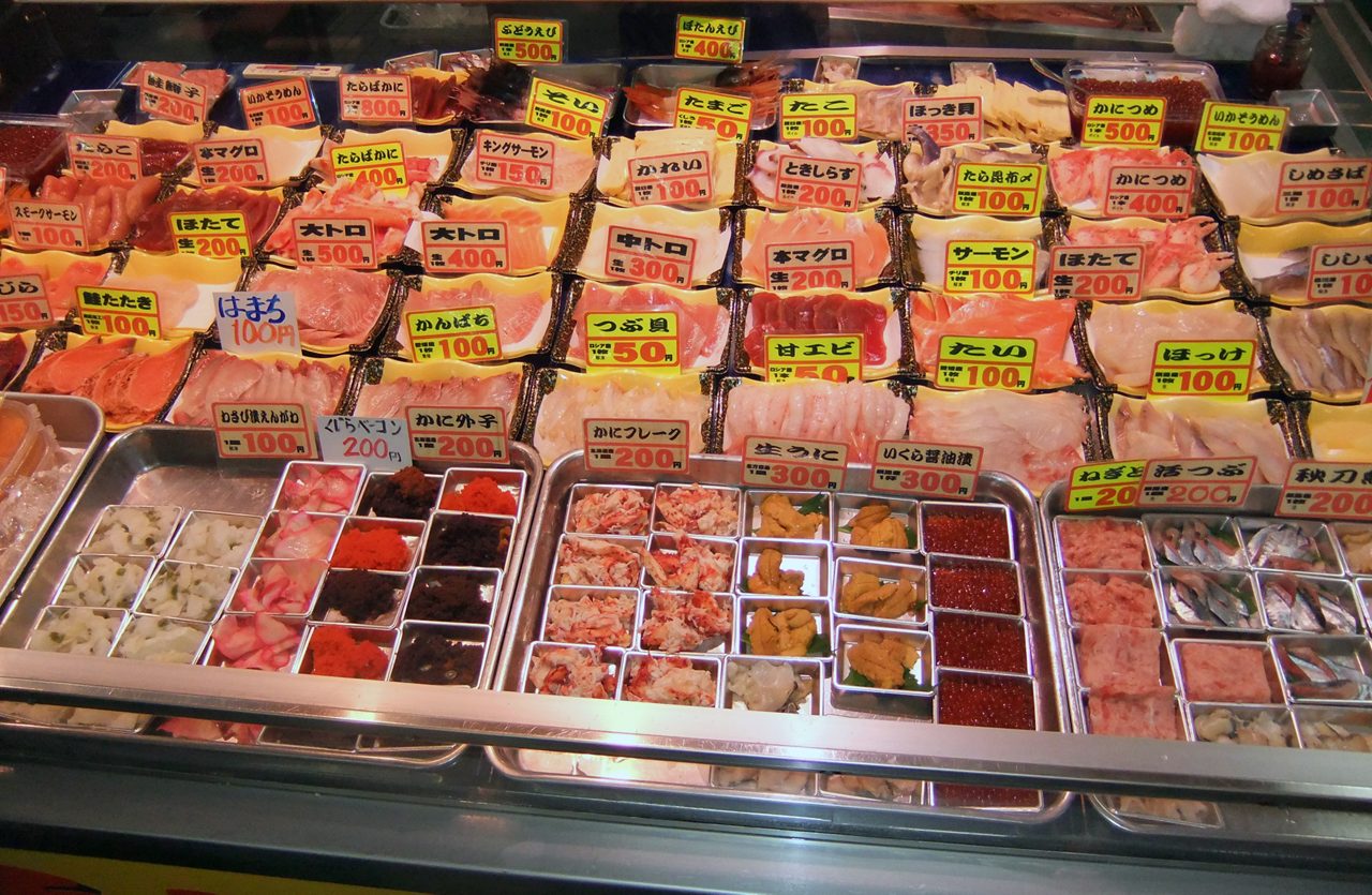 Close-up of a seafood stall in Kushiro Market displaying sashimi slices, fish roe and other toppings with handwritten price signs.