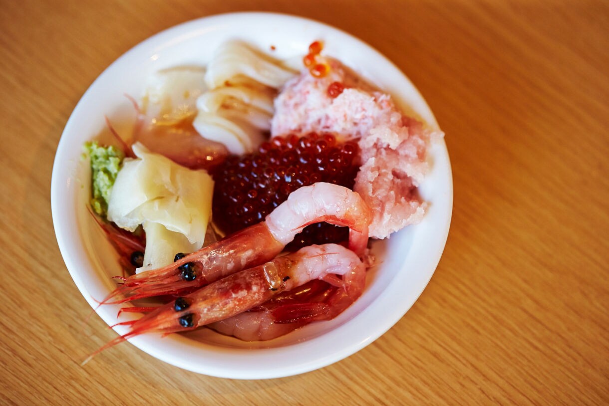 A bowl of assorted seafood including raw shrimp, salmon roe, crab, squid, wasabi and ginger on a wooden table.